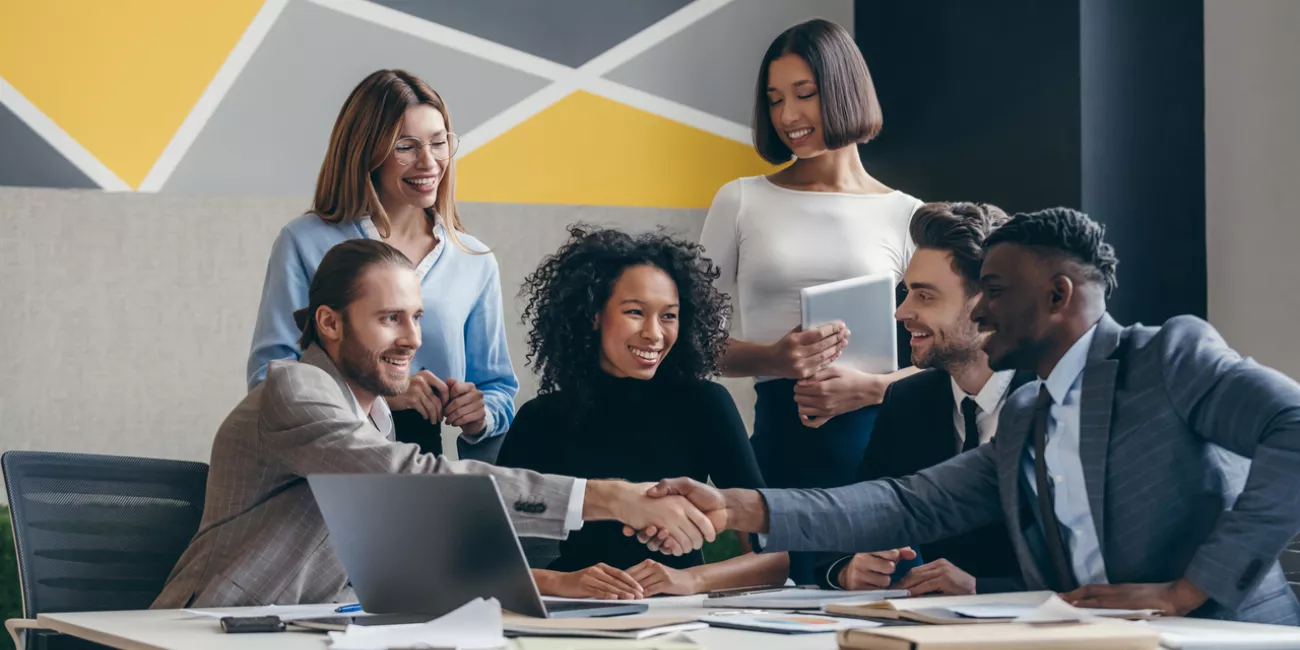Two smiling young businessmen shaking hands while sitting with their colleagues at the office desk
