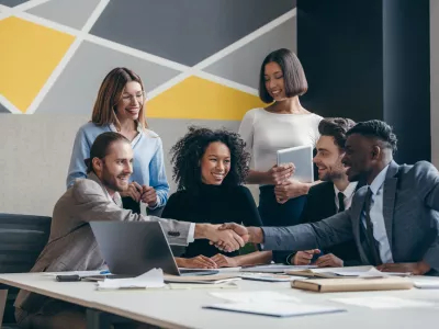 Two smiling young businessmen shaking hands while sitting with their colleagues at the office desk