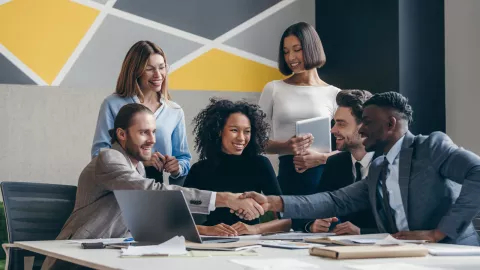 Two smiling young businessmen shaking hands while sitting with their colleagues at the office desk